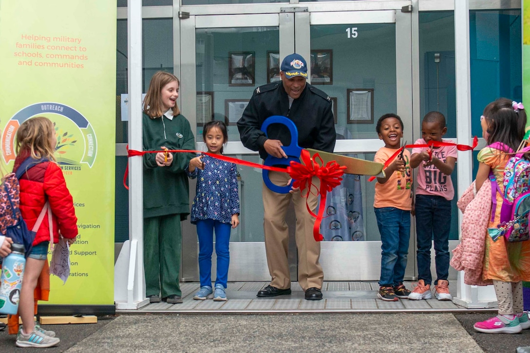 A Navy officer and a group of children use scissors to cut a red ribbon.