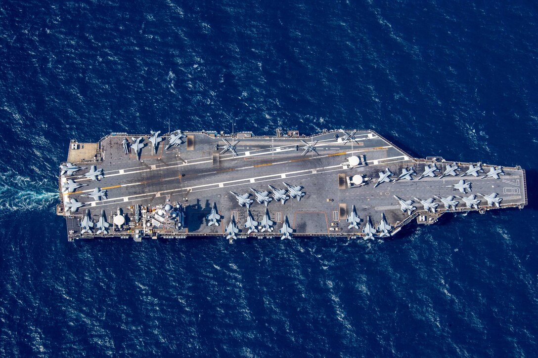 An aerial view of an aircraft carrier moving through dark blue water.