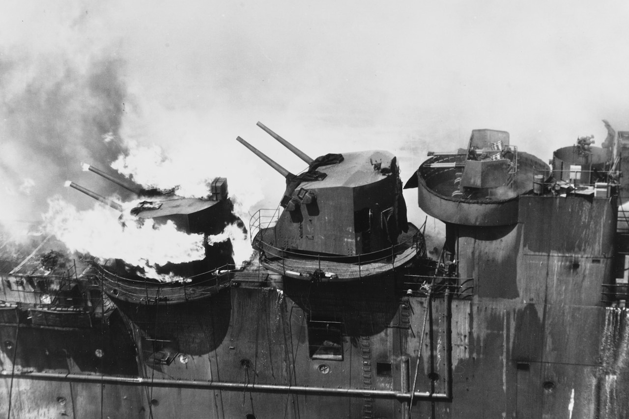 Flames shoot from one of two gun mounts on a large ship.