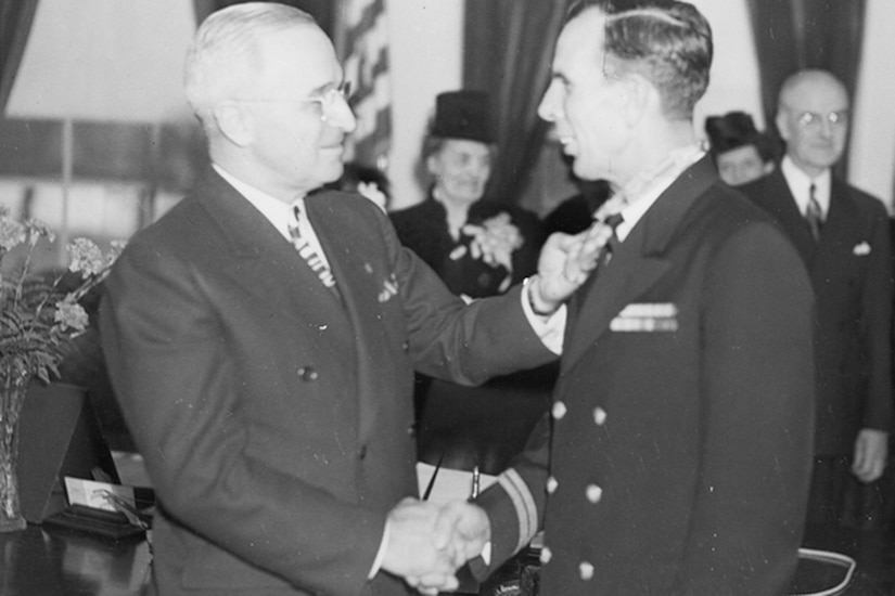 Two men shake hands. One touches a medal on the other man’s neck.
