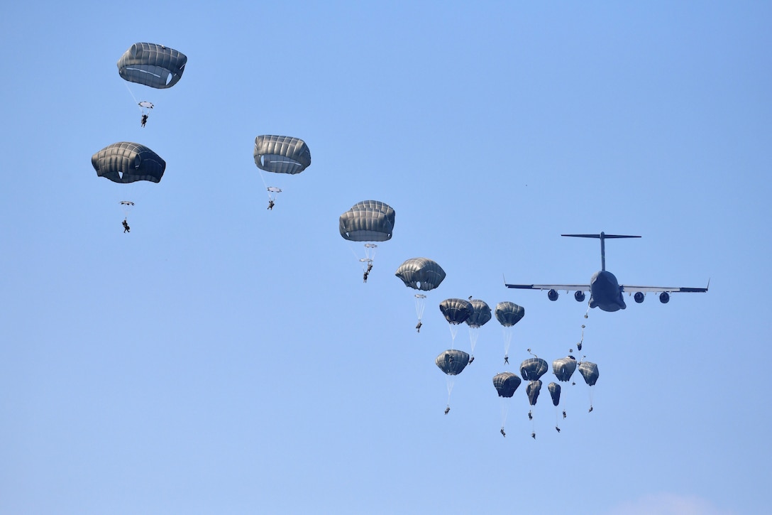 Soldiers free fall with parachutes as an aircraft flies in the background.