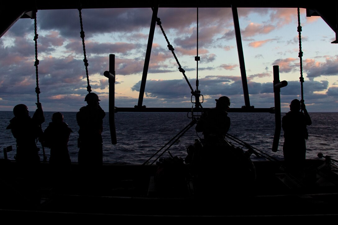 A group of sailors in silhouette face the ocean and dark cloudy sky as they pull on lines from above on a ship's deck.