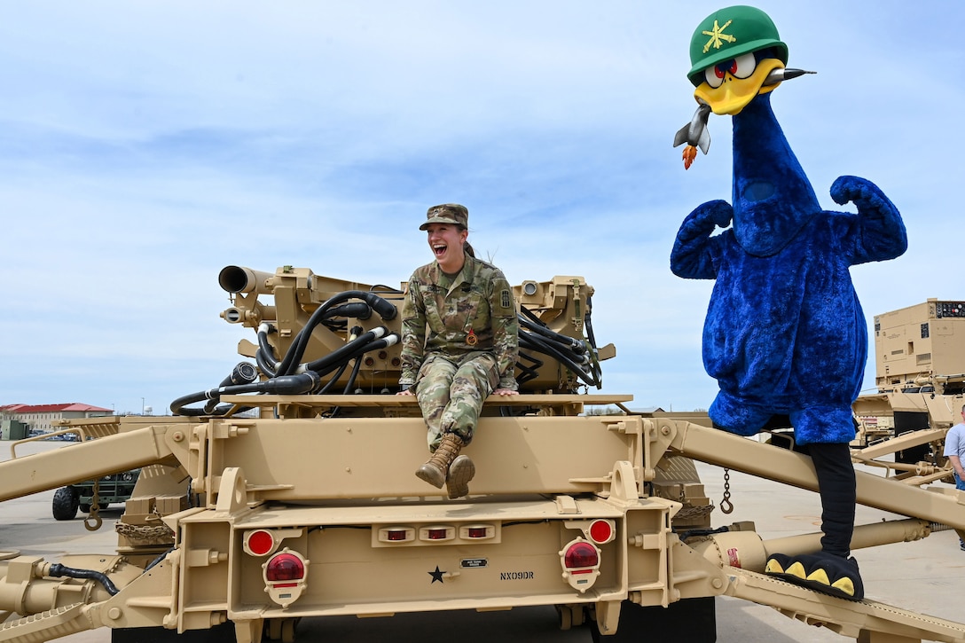 A soldier sitting on a vehicle laughs as someone dressed as a big blue bird stands next to her.