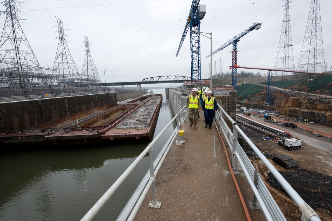 Lt. Col. Joseph Sahl, U.S. Army Corps of Engineers Nashville District commander, leads a group of stakeholders on a tour of Kentucky Lock and the Kentucky Lock Addition Project March 22, 2023, on the Tennessee River in Grand Rivers, Kentucky. He is having a conversation with Cmdr. Jennifer Andrew, commander of U.S. Coast Guard Marine Safety Unit Paducah, as the group walked on the coffer dam and future downstream approach wall during the tour. A vessel and barges approach the downstream active lock on the left of the photo and construction of the new lock chamber can be seen on the right of the photo. (USACE Photo by Lee Roberts)