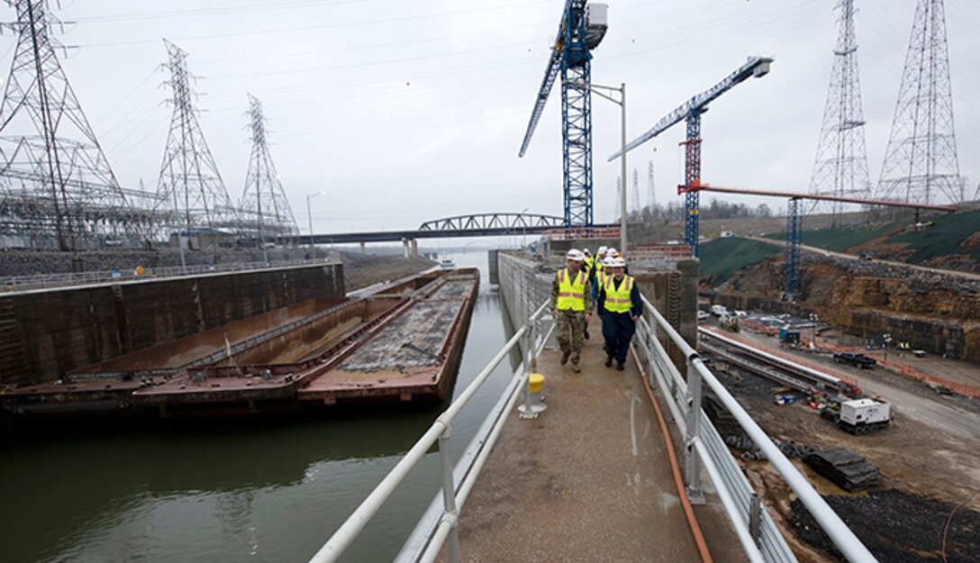 Lt. Col. Joseph Sahl, U.S. Army Corps of Engineers Nashville District commander, leads a group of stakeholders on a tour of Kentucky Lock and the Kentucky Lock Addition Project March 22, 2023, on the Tennessee River in Grand Rivers, Kentucky. He is having a conversation with Cmdr. Jennifer Andrew, commander of U.S. Coast Guard Marine Safety Unit Paducah, as the group walked on the coffer dam and future downstream approach wall during the tour. A vessel and barges approach the downstream active lock on the left of the photo and construction of the new lock chamber can be seen on the right of the photo. (USACE Photo by Lee Roberts)