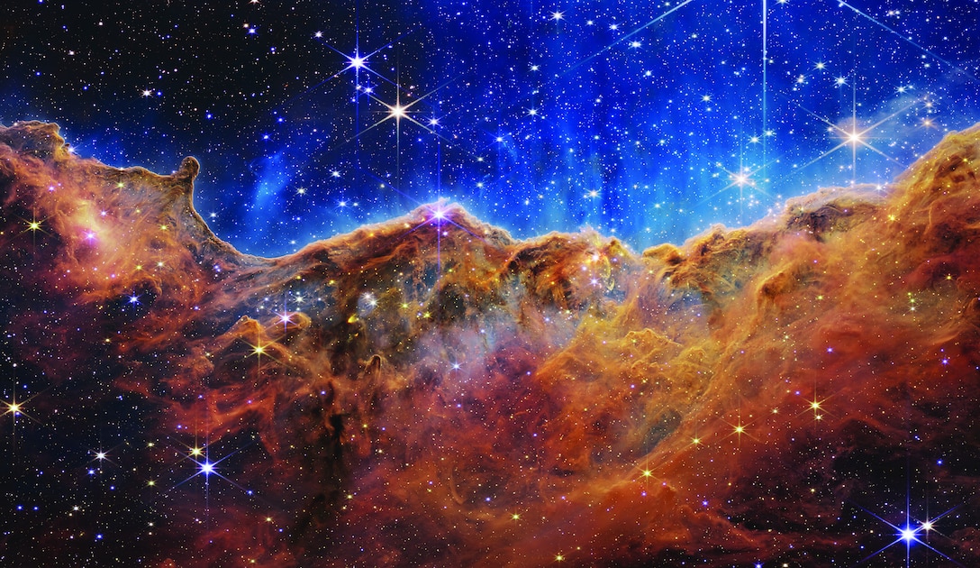 What looks much like craggy mountains on a moonlit evening is actually the edge of a nearby, young, star-forming region NGC 3324 in the Carina Nebula. Captured in infrared light by the Near-Infrared Camera (NIRCam) on NASA’s James Webb Space Telescope, this image reveals previously obscured areas of star birth.
Called the Cosmic Cliffs, the region is actually the edge of a gigantic, gaseous cavity within NGC 3324, roughly 7,600 light-years away. The cavernous area has been carved from the nebula by the intense ultraviolet radiation and stellar winds from extremely massive, hot, young stars located in the center of the bubble, above the area shown in this image. The high-energy radiation from these stars is sculpting the nebula’s wall by slowly eroding it away.  
NIRCam – with its crisp resolution and unparalleled sensitivity – unveils hundreds of previously hidden stars, and even numerous background galaxies. Several prominent features in this image are described below.
•	The “steam” that appears to rise from the celestial “mountains” is actually hot, ionized gas and hot dust streaming away from the nebula due to intense, ultraviolet radiation. 
•	Dramatic pillars rise above the glowing wall of gas, resisting the blistering ultraviolet radiation from the young stars.
•	Bubbles and cavities are being blown by the intense radiation and stellar winds of newborn stars.
•	Protostellar jets and outflows, which appear in gold, shoot from dust-enshrouded, nascent stars.
•	A “blow-out” erupts at the top-center of the ridge, spewing gas and dust into the interstellar medium. 
•	An unusual “arch” appears, looking like a bent-over cylinder.
This period of very early star formation is difficult to capture because, for an individual star, it lasts only about 50,000 to 100,000 years – but Webb’s extreme sensitivity and exquisite spatial resolution have chronicled this rare event.
Located roughly 7,600 light-years away, NGC 3324 was first cat