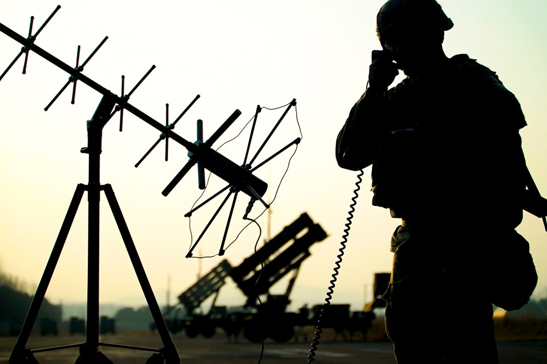 A soldiers speaks on the phone next to an antenna as shown in silhouette.