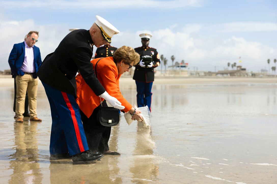 A retired Marine supports a woman as she pours ashes into a body of water while Marines and family watch.