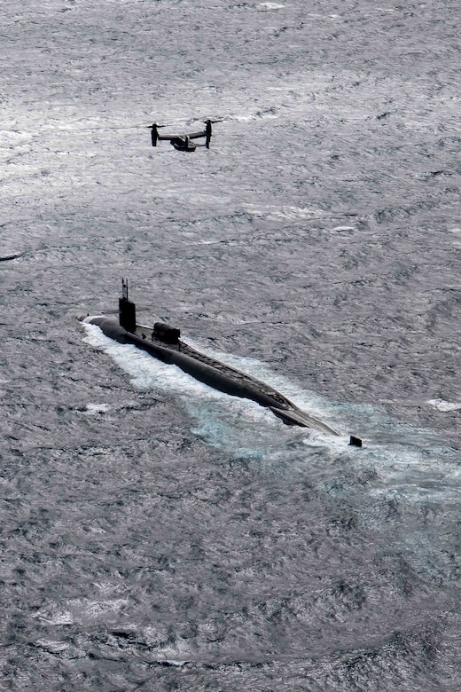 An aircraft flies over a submarine transiting a body of water.