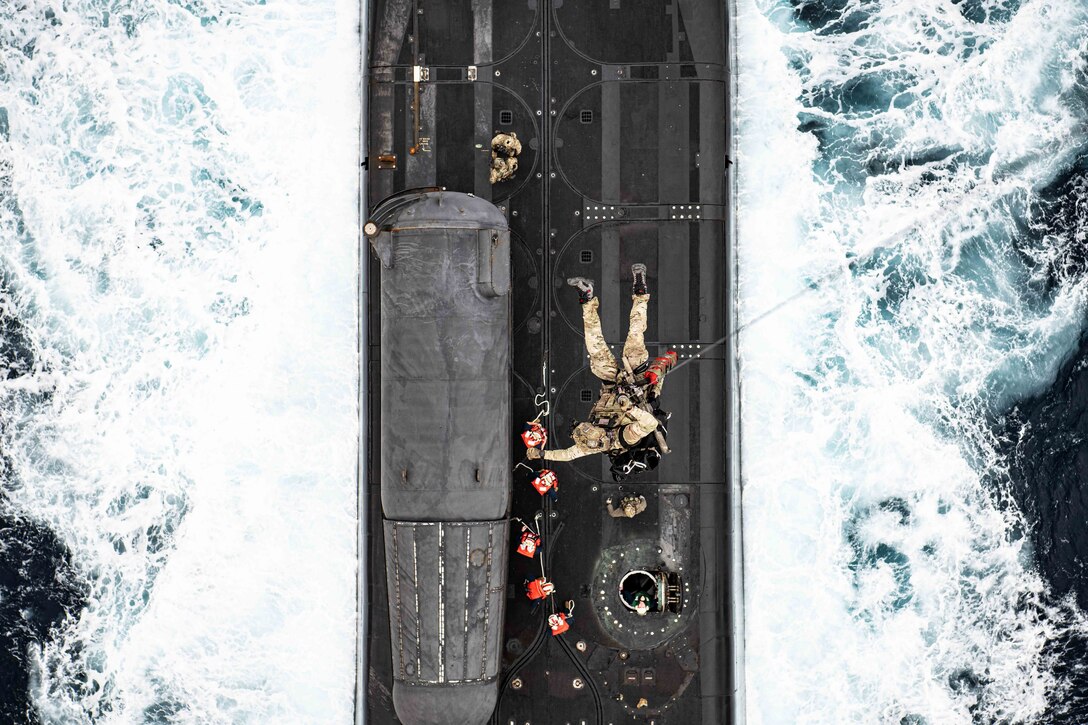 Sailors attached to helicopter by rope rappel onto a ship at sea.