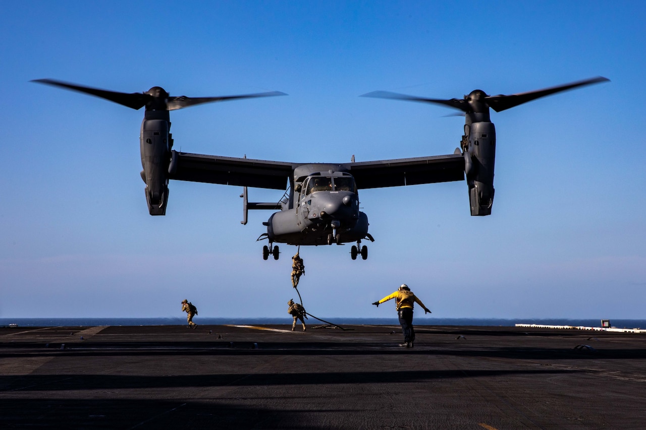 Navy SEALs rappel down a rope attached to a helicopter on to a ship at sea.