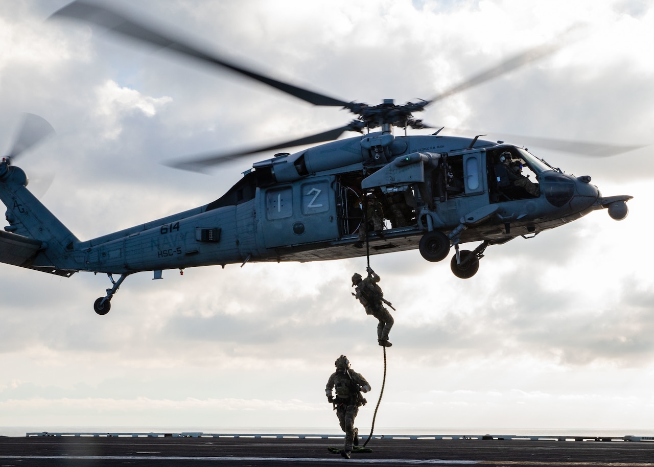 Two sailors rappel down a rope attached to a helicopter on to a ship at sea.