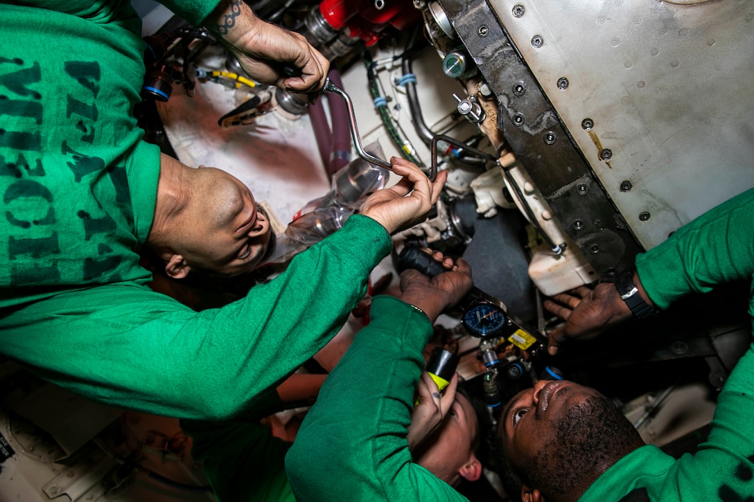 Three sailors perform maintenance on ship equipment.