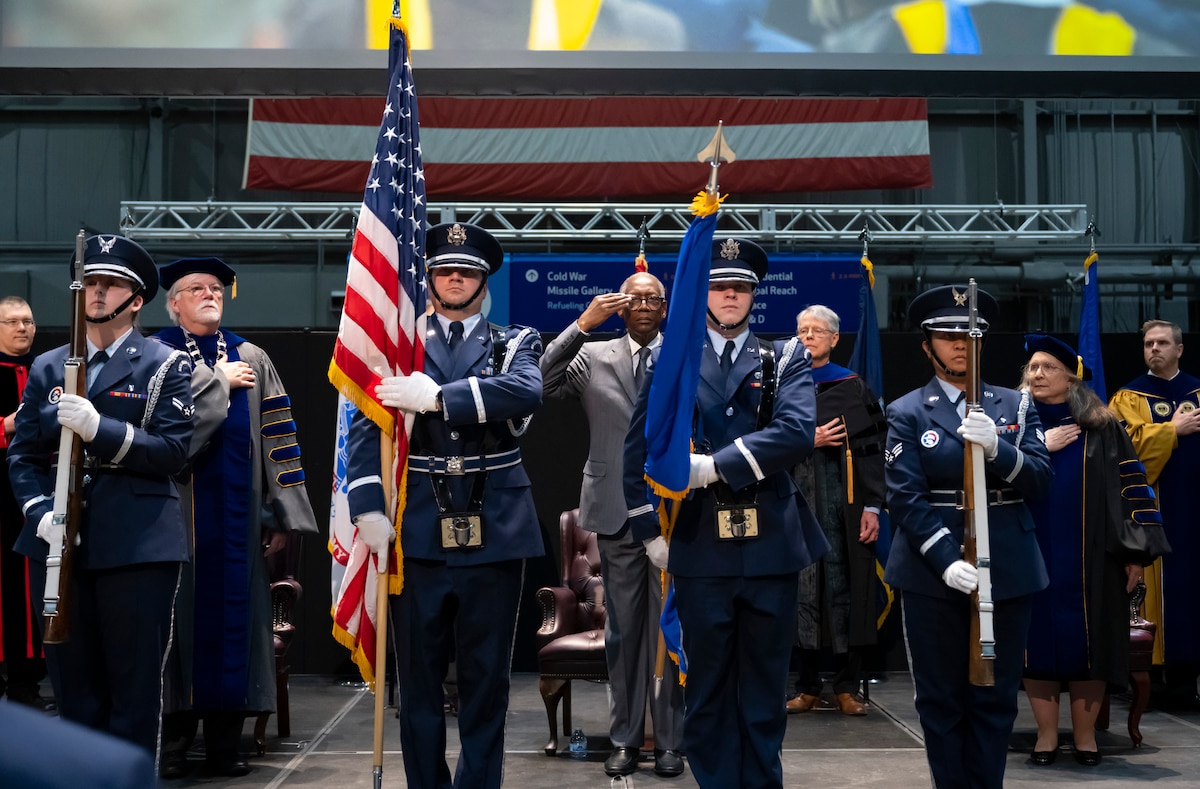 Four Airmen in dress uniform stand at attention holding rifles and flags. Behind them, either saluting or with their hands on their hearts, are people in academic regalia. The man in the center is in a civilian suit.