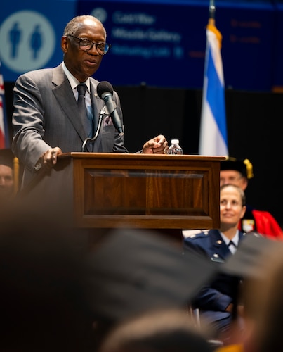African-American man in civilian clothes speaks at a podium. The heads of some of the audience can be seen in the foreground.