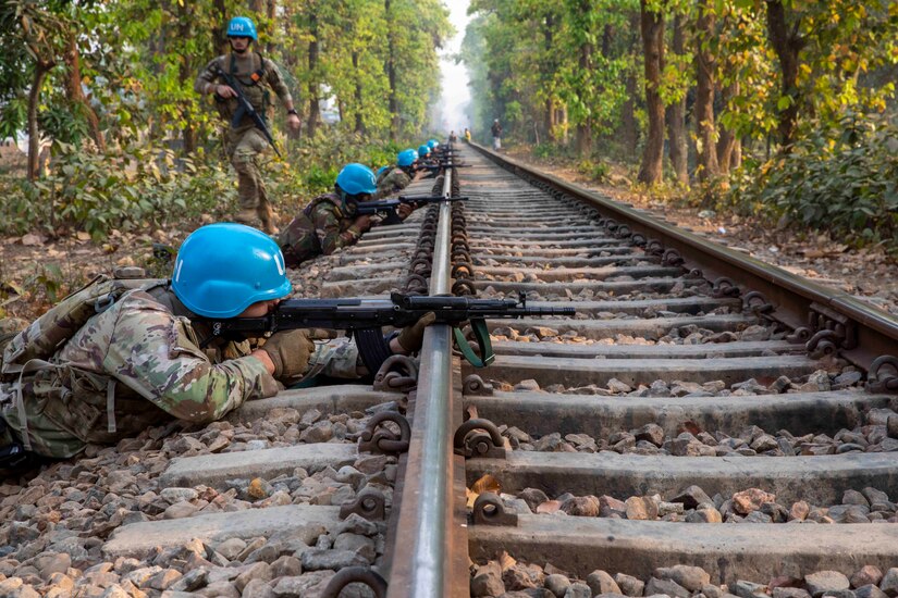 Guardsmen lie in a line across train tracks while holding weapons as another guardsman walks behind them.