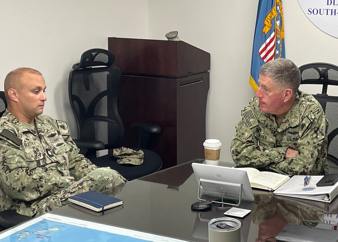 Two Navy officers discuss sit at a table for a discussion.