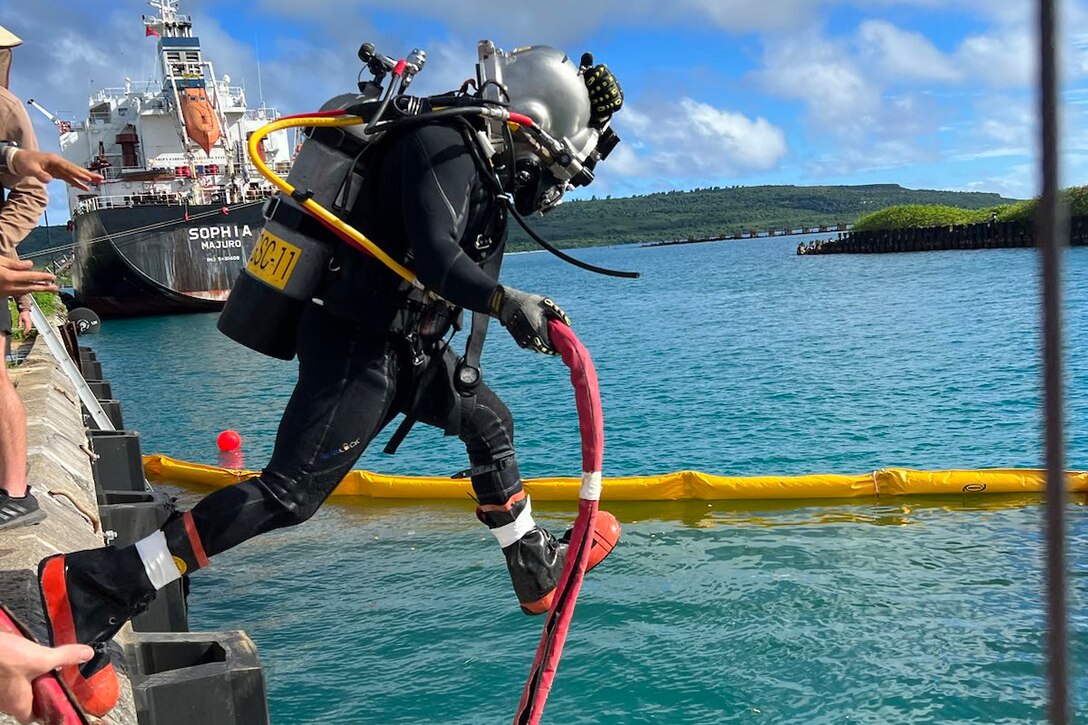 A sailor wearing diving gear jumps into a body of water with a ship in the background.