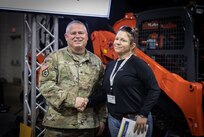 BG Gene Holt, a smiling white man in military camo uniform, shakes the hand of a brunette white woman, Maryann Perry