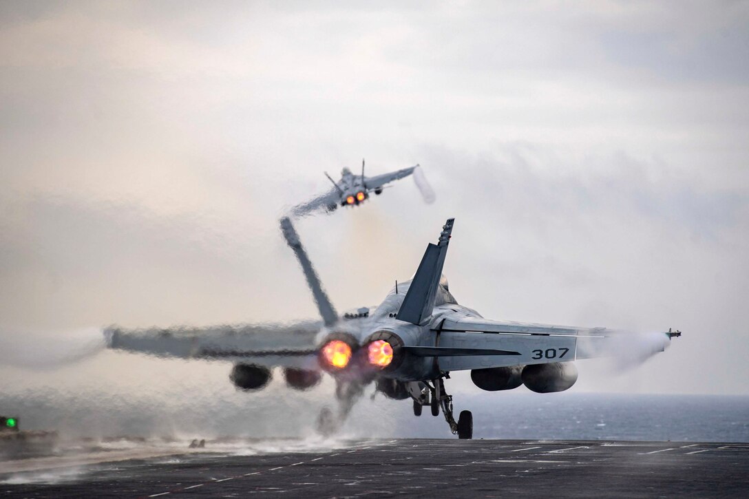 An aircraft takes off from the flight deck of a ship as another aircraft flies above.