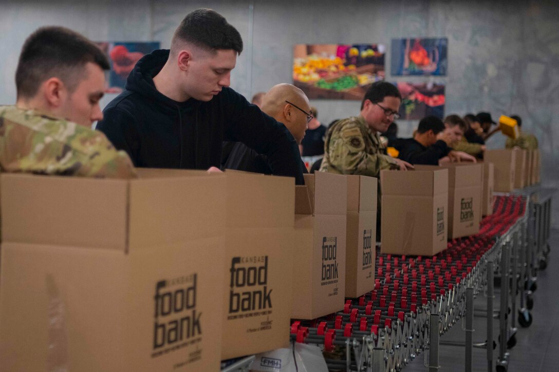 Airmen stand on a production line filling boxes with canned goods.