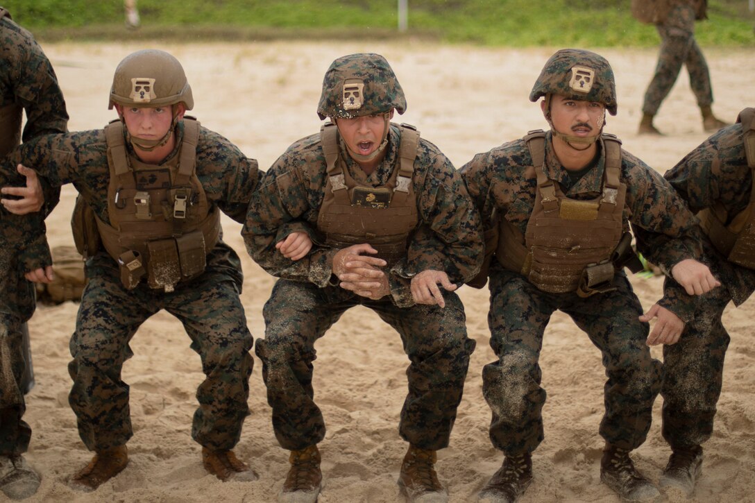 Marines lock arms next to each other while squatting on the sand.