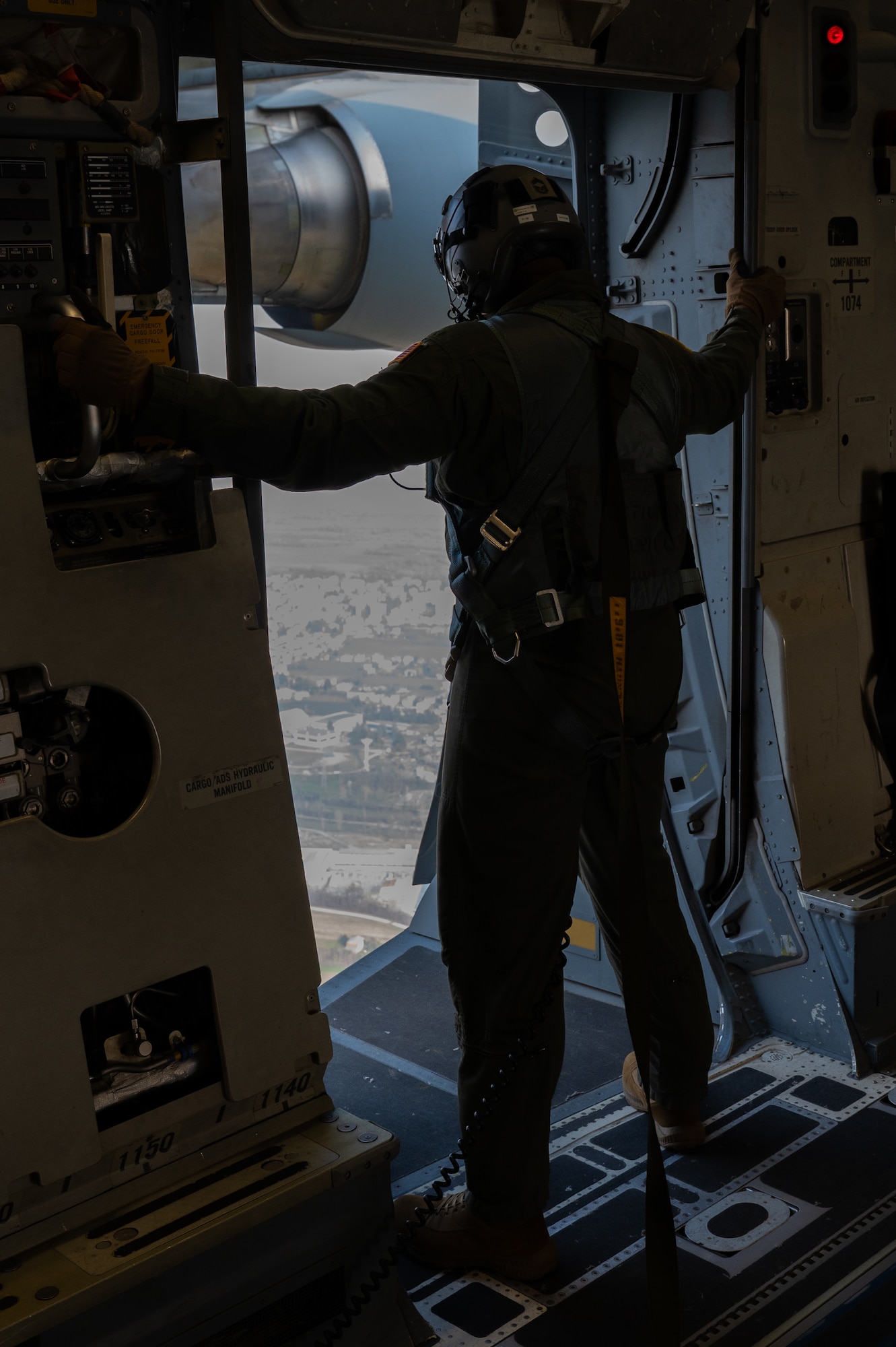 A loadmaster stands in full gear in front of an open door as a C17 flies across Italy.