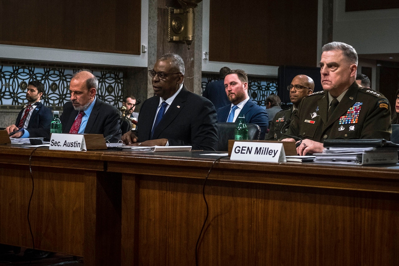 Three men are pictured seated behind a desk with others in rows behind them.