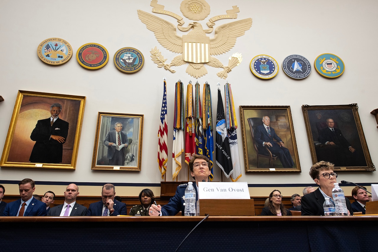 Two service members sit at a table in front of microphones with others sitting  behind.