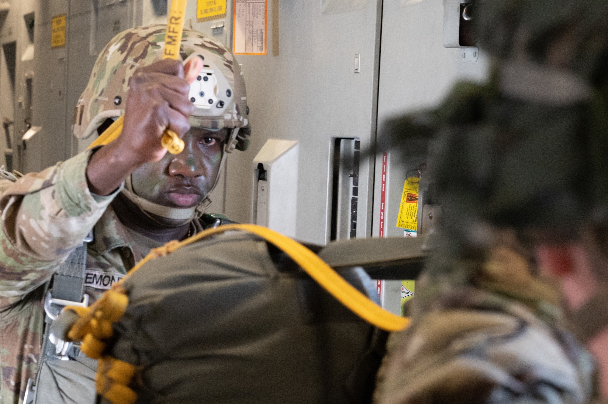 A U.S. Army soldier in full camo face paint and parachute gear stands by waiting his turn to jump while holding a yellow strap.