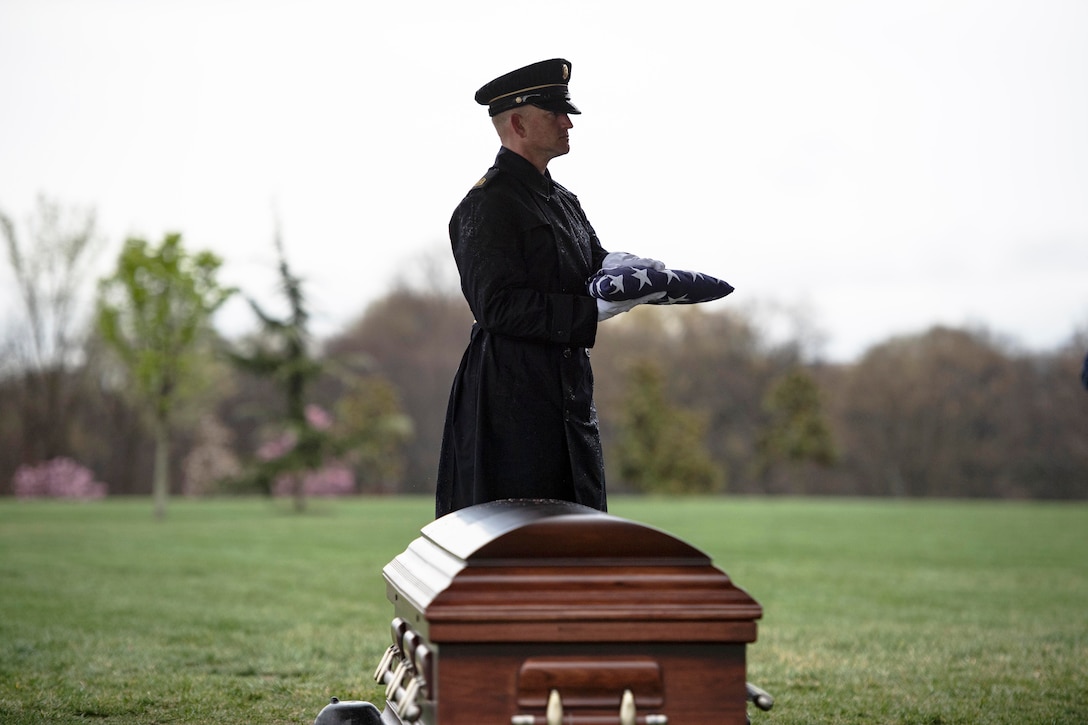 A soldier stands behind a casket holding a folded American flag.