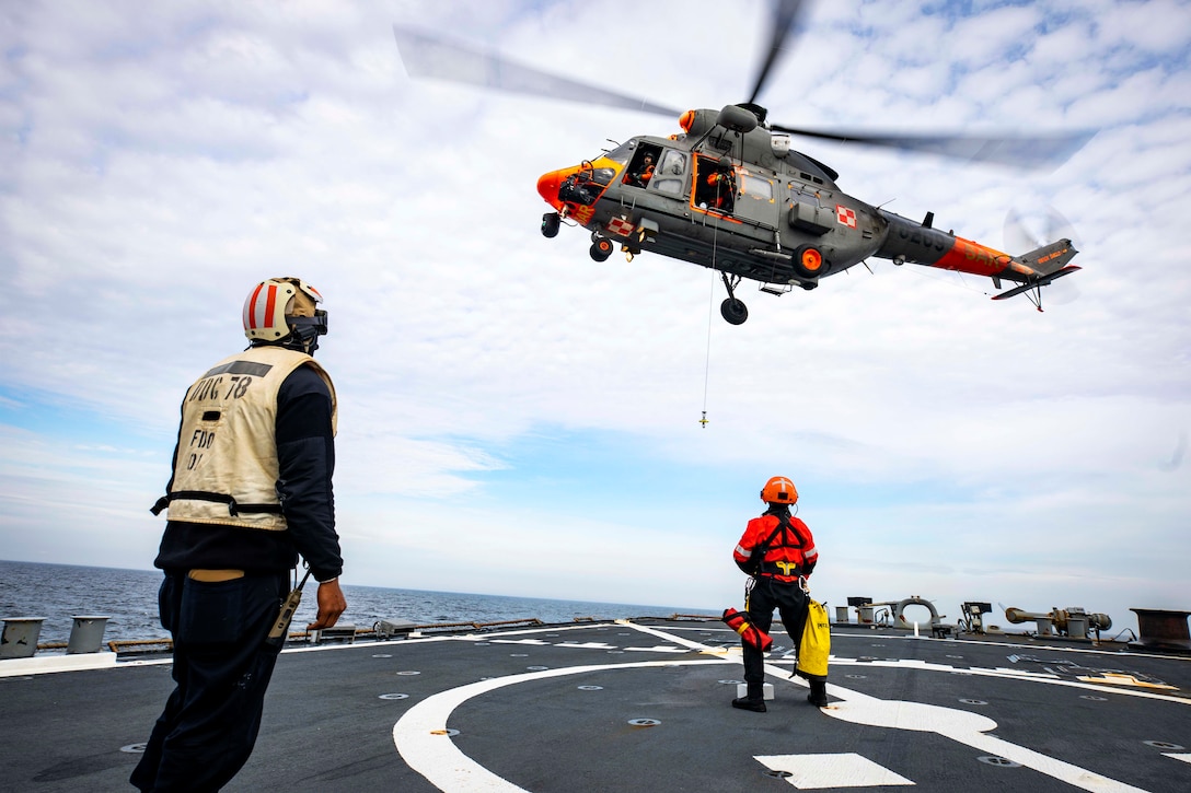 A  helicopter hovers above two sailors on the flight deck of a ship.