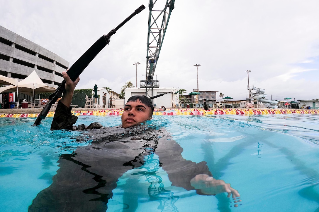 A uniformed Marine swims while holding a rifle in the foreground as two others swim in the background.