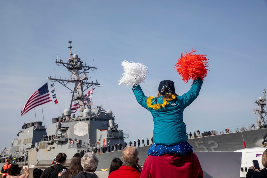 A child sits on a person’s shoulders as she waves pompoms with a Navy ship in the distance.