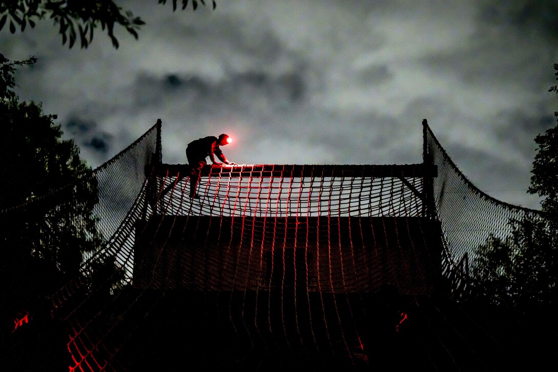 An airman wearing a search light crawls across a log covered by a net in the dark illuminated by a red light.