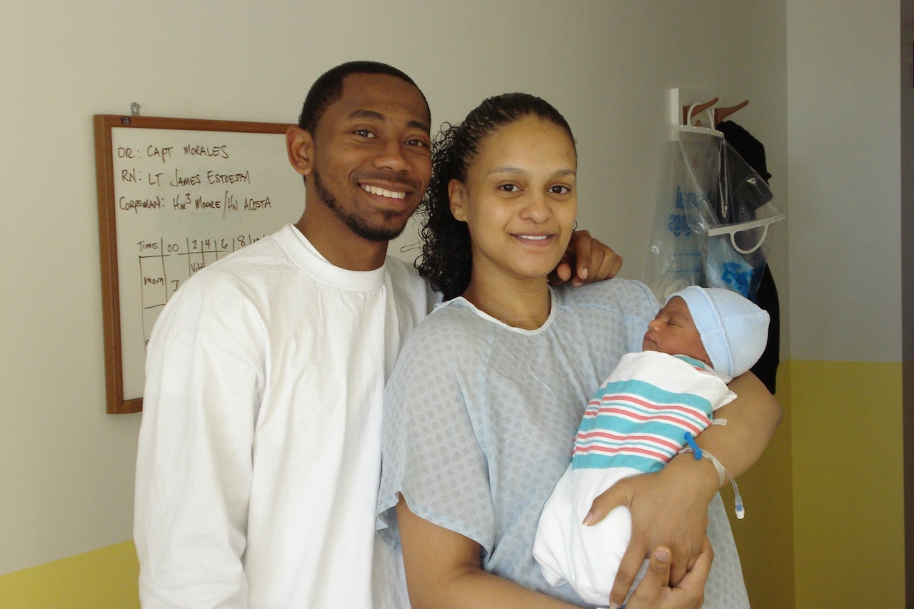 A man stands with his arm on the shoulder of a woman dressed in a hospital gown; in her arms is a newborn baby swaddled in a blanket.
