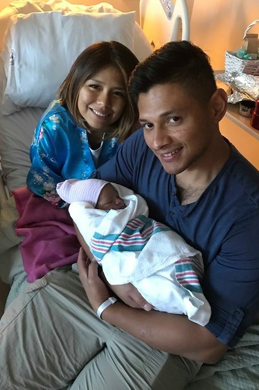 A woman in a hospital gown looks over the shoulder of a man as he cradles an infant in his arms.