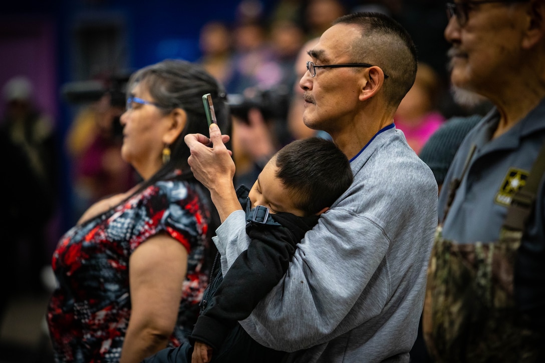Jerome Apatiki, while holding one of his nine grandchildren, takes video during the Alaska Heroism Medal ceremony March 28, 2023, at the John Apangalook Memorial High School gymnasium in Gambell, Alaska. Apatiki received the medal on behalf of Pfc. Holden Apatiki Sr. for his actions during a 1955 rescue mission after two Russian MiG-15s shot down a U.S. Navy P2V-5 Neptune plane flying a routine maritime patrol over the Bering Sea with 11 sailors on board.