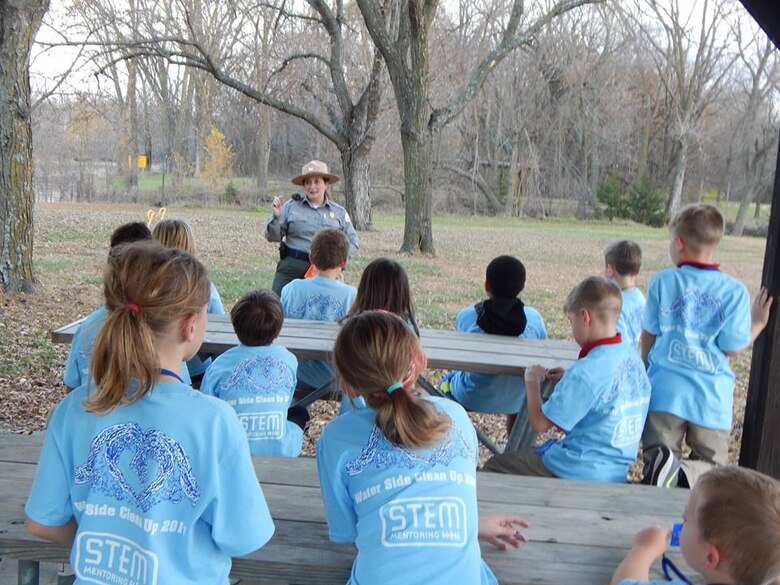 A woman in a park ranger uniform talks to a group of children in blue shirts outside.