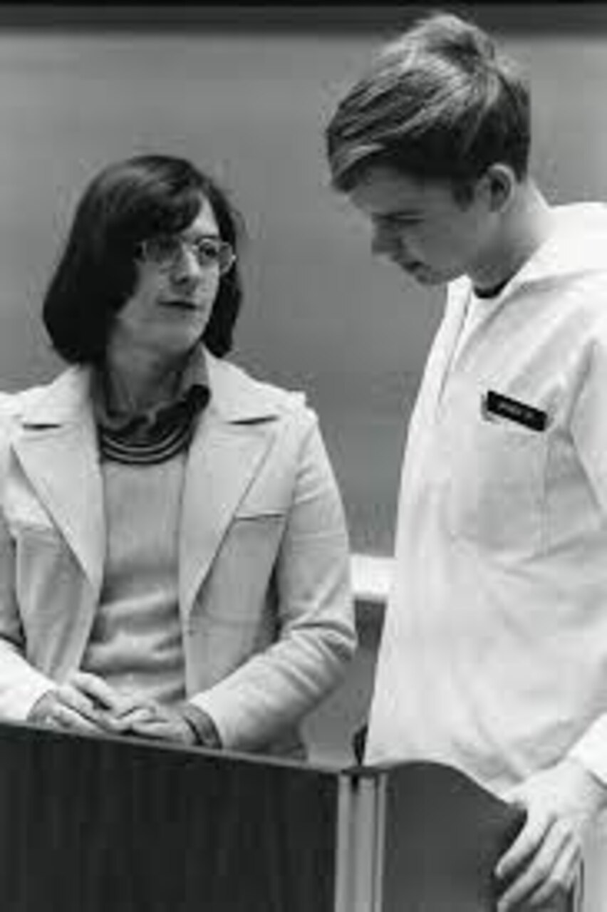 Black and white photo of Rae Jean Goodman, standing on left talking to a young man on the right