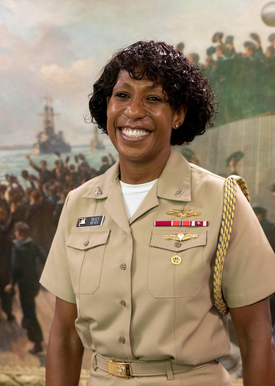 Smiling woman in beige uniform posing for camera with historic navy wall behind her.