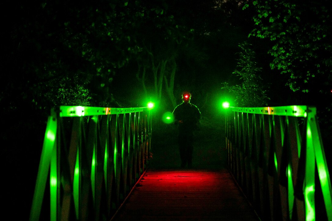 An airman walks across a bridge illuminated by green light at night.