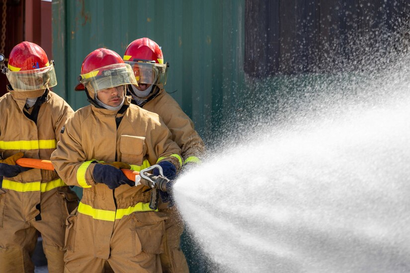 Sailors in firefighting gear shoot water from a hose.