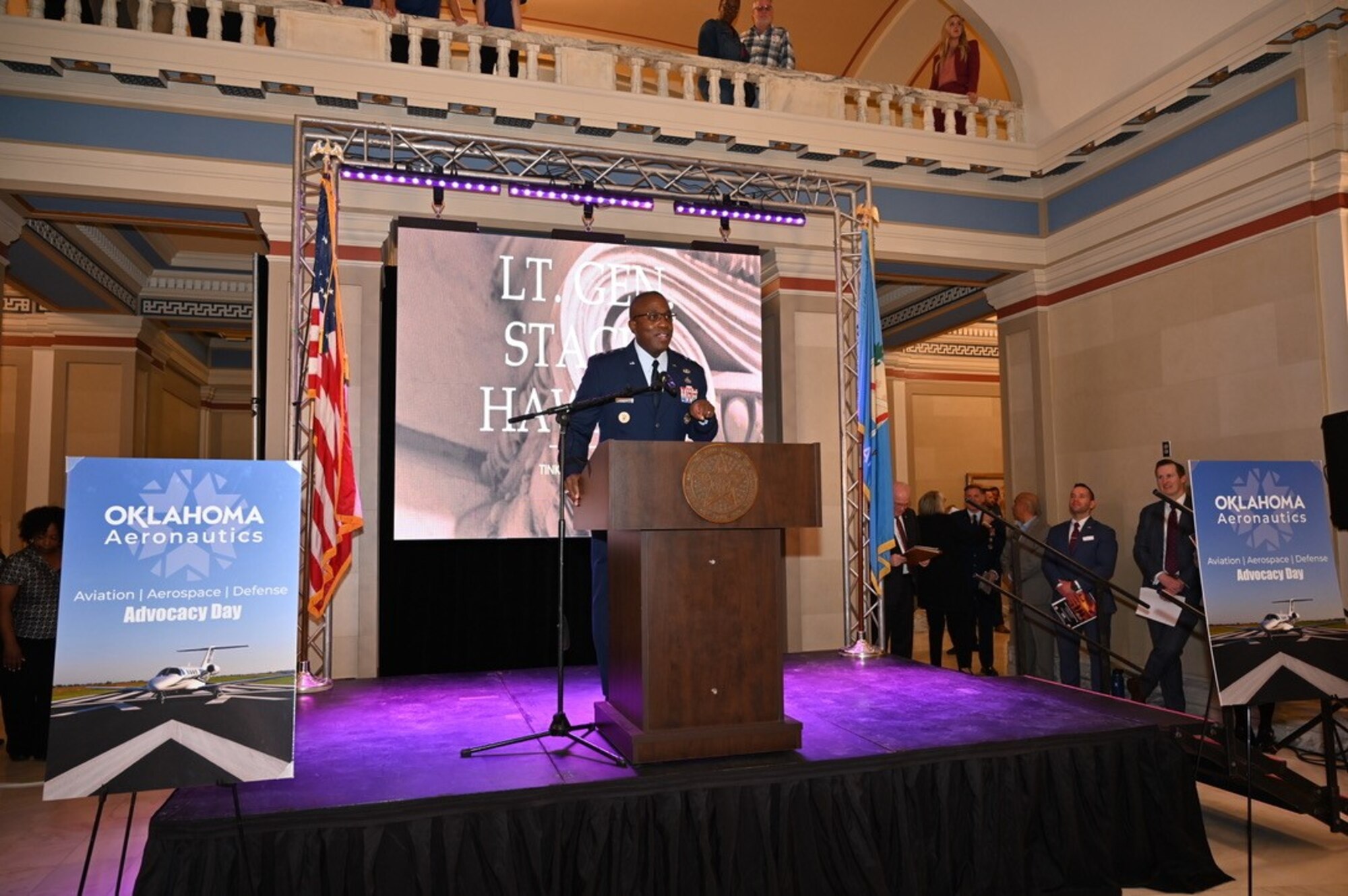 Lt Gen Hawkins stands at a podium in front of a large electronic screen with his name on it.