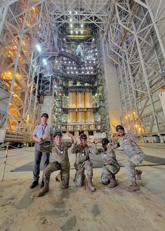 Four uniformed troops gesture to raise a roof while wearing hardhats; civilian stands by them.