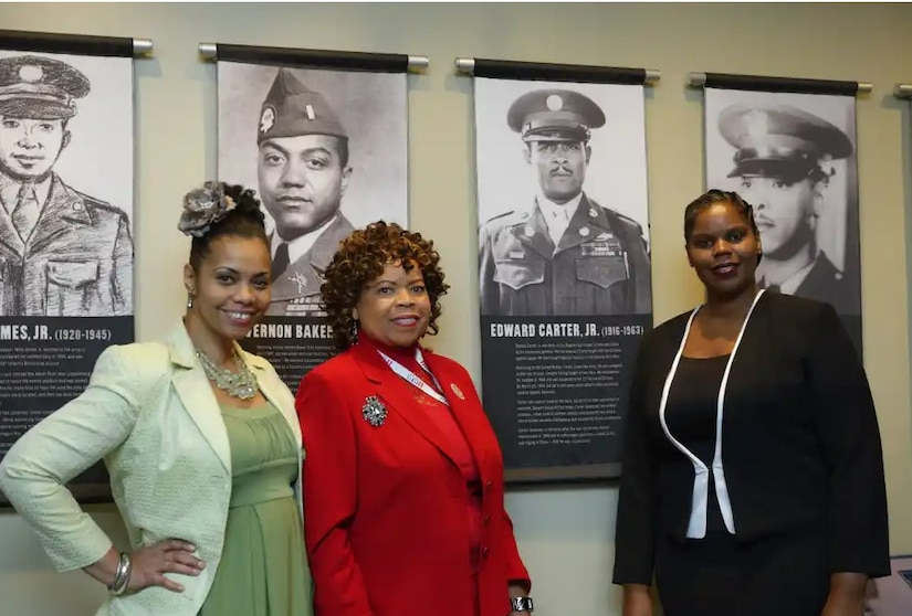 Three women pose in front of a banner bearing the image of a service member.