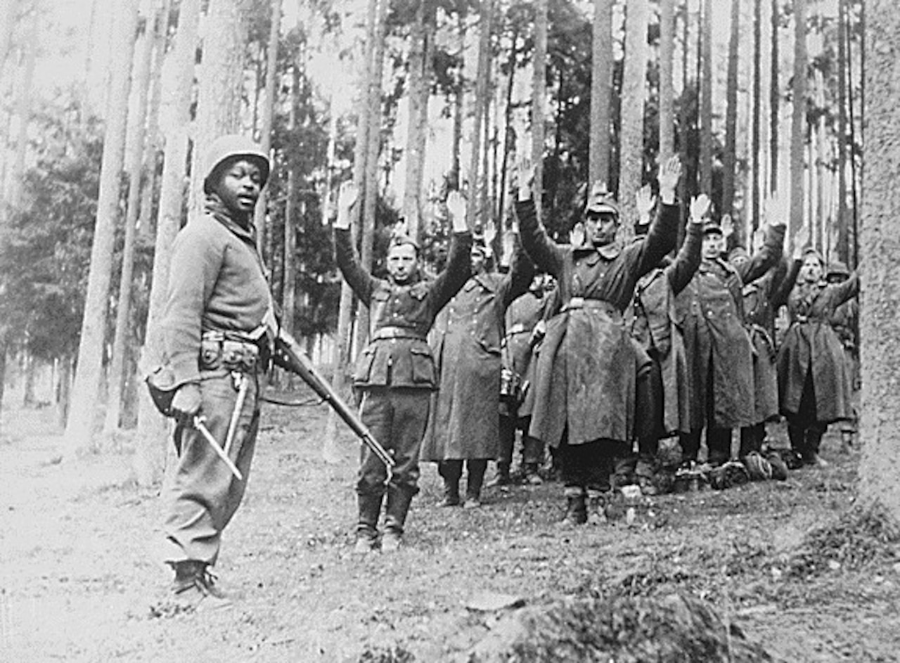 A man with a rifle stands in front of several men who have their hands in the air in surrender.
