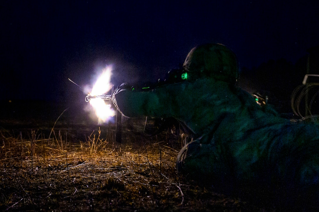 A soldier laying on the ground in the dark fires a weapon illuminated by green and white lights.