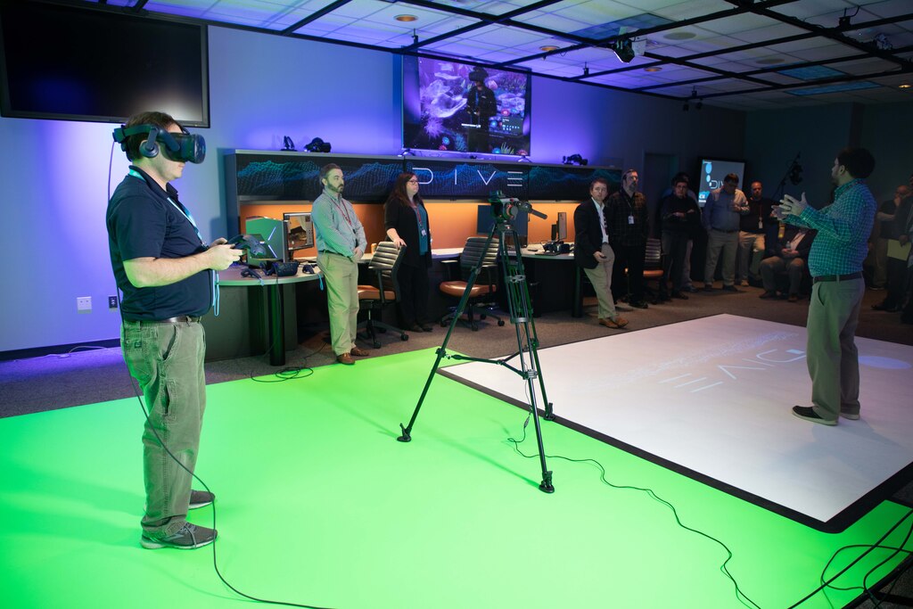 A man stands in the foreground with a virtual reality set on his face and holds a controller while standing on a green screen. Several people are in the background watching.