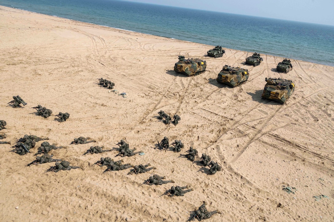U.S. and South Korean marines kneel with weapons on the sand while fellow service members are parked behind them in armored vehicles with the beach in the background.